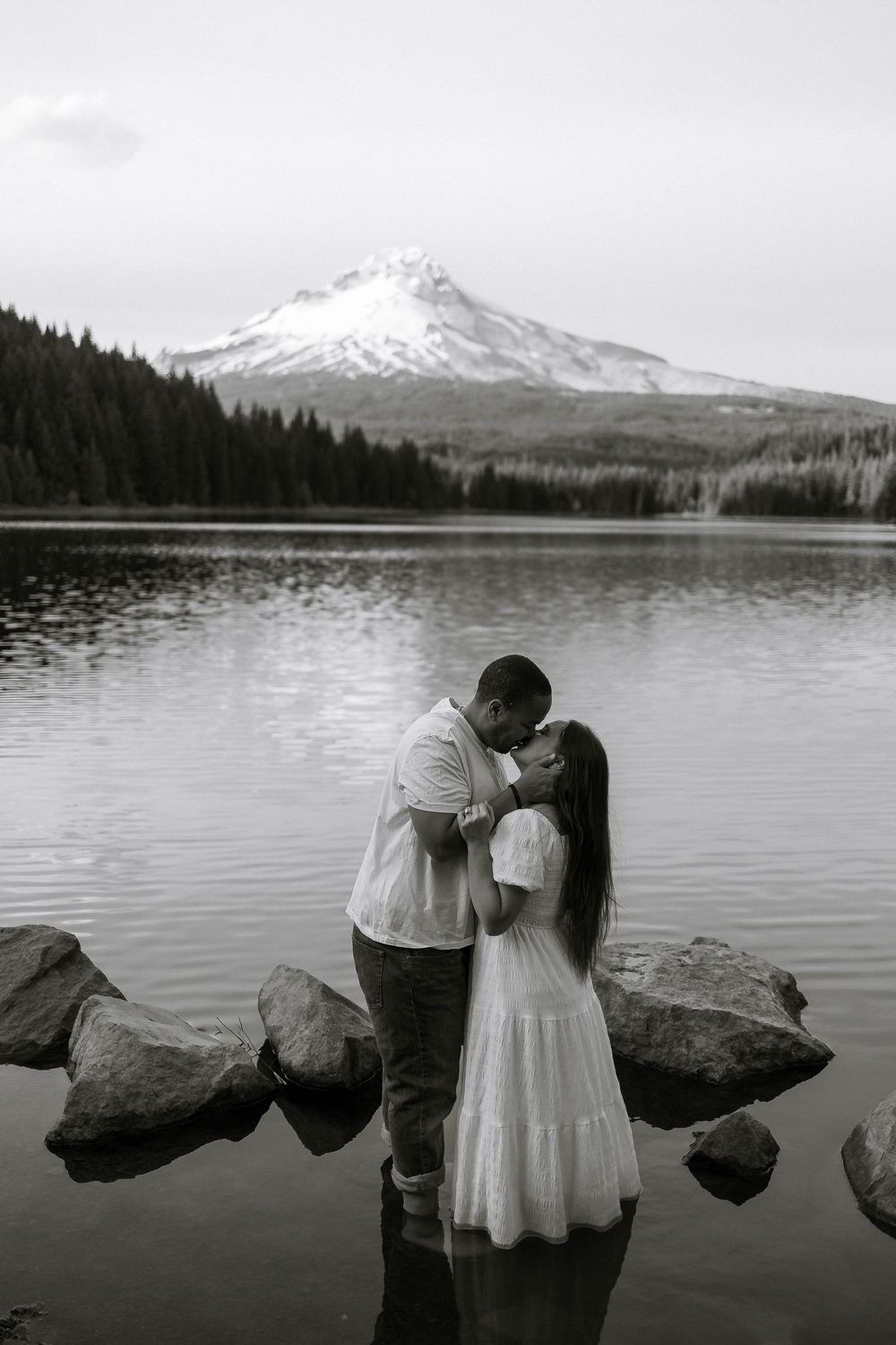 Couple kissing at Trillium Lake with snow-capped Mount Hood reflecting in water, black and white engagement photo.
