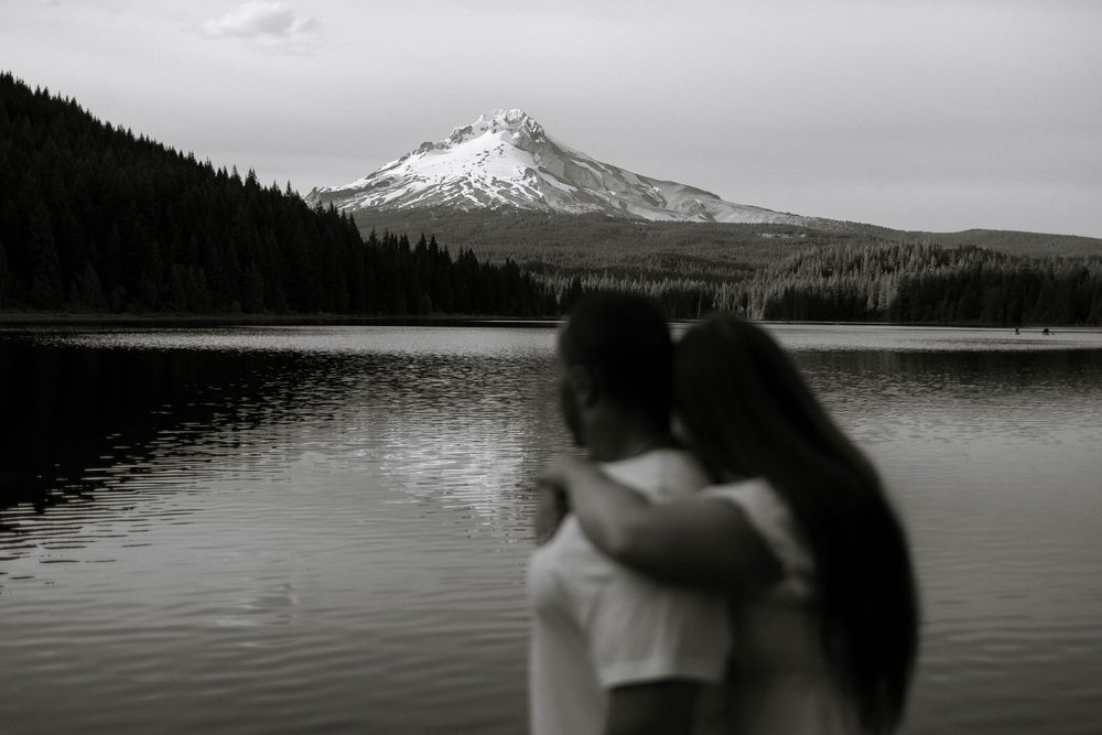 Black and white photo of a couple embracing by a lake with Mount Hood reflected in calm water.