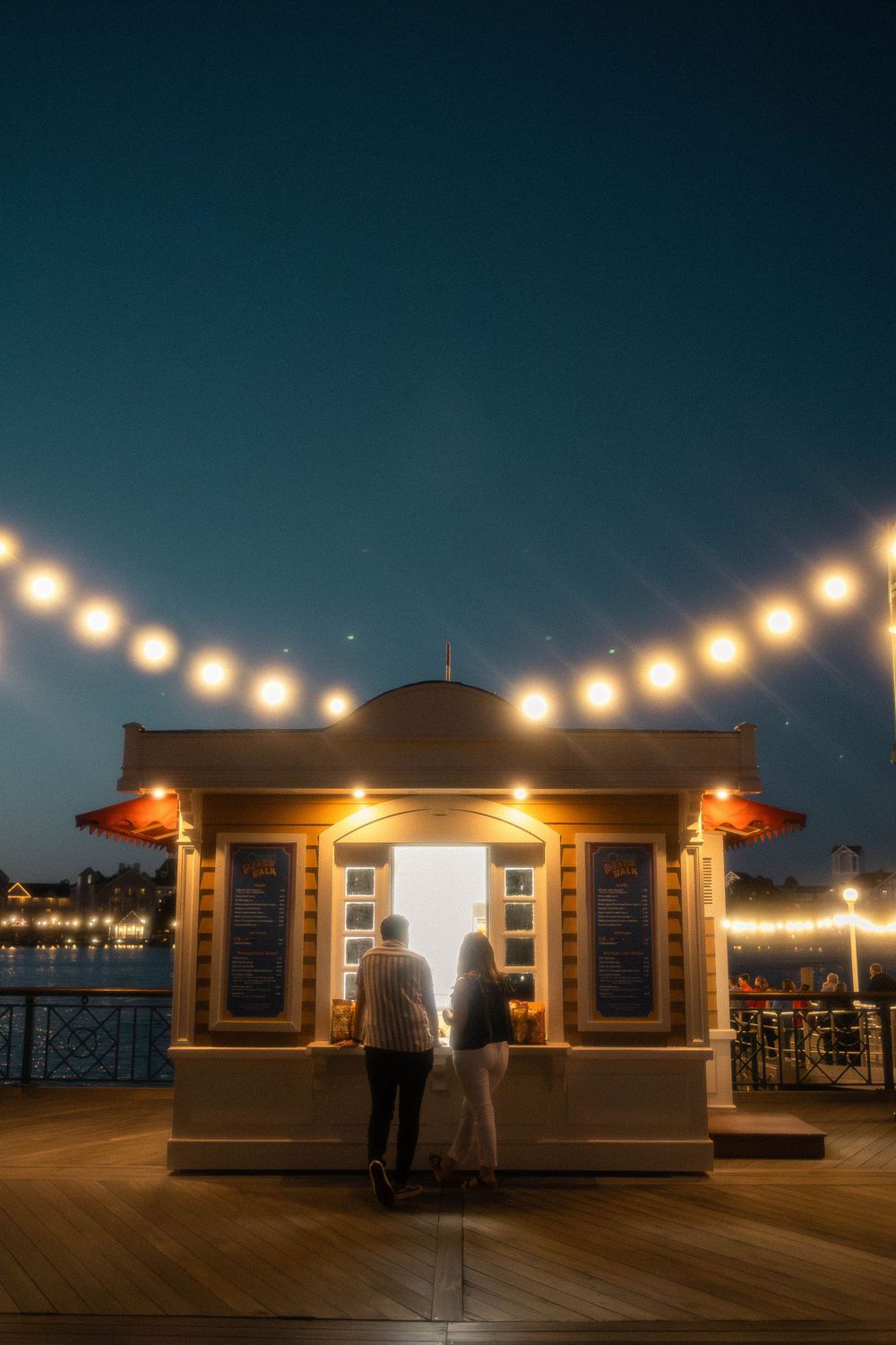 String lights illuminate a wooden pier deck with small beach huts at night creating a cozy coastal atmosphere.