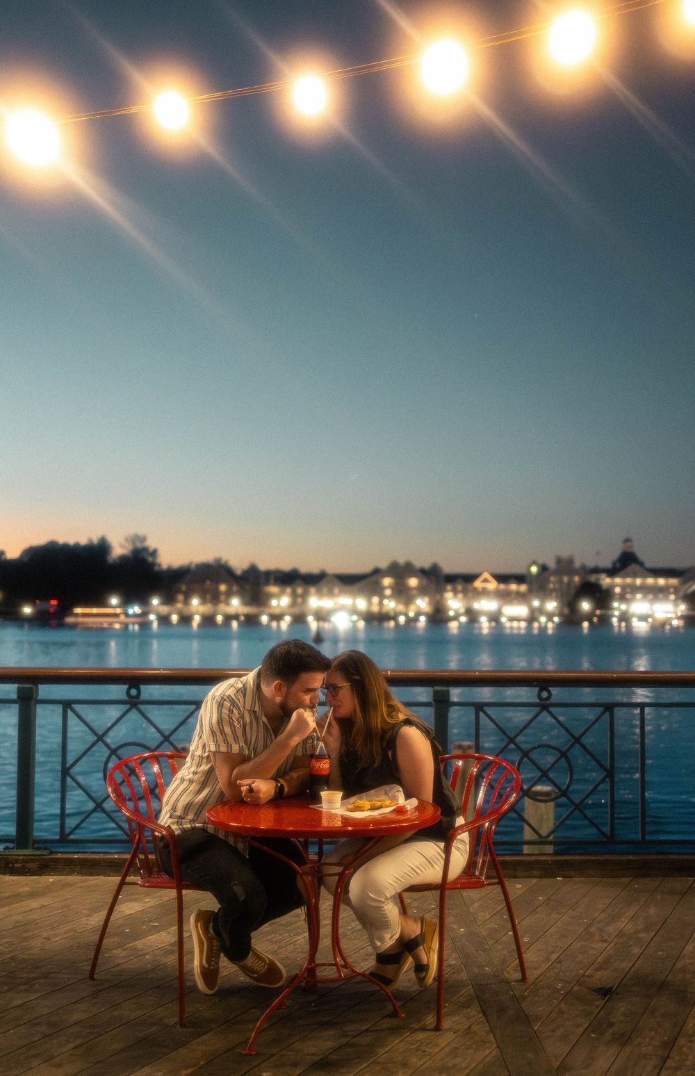 Couple sharing a romantic dinner at a waterfront cafe table with string lights at dusk.