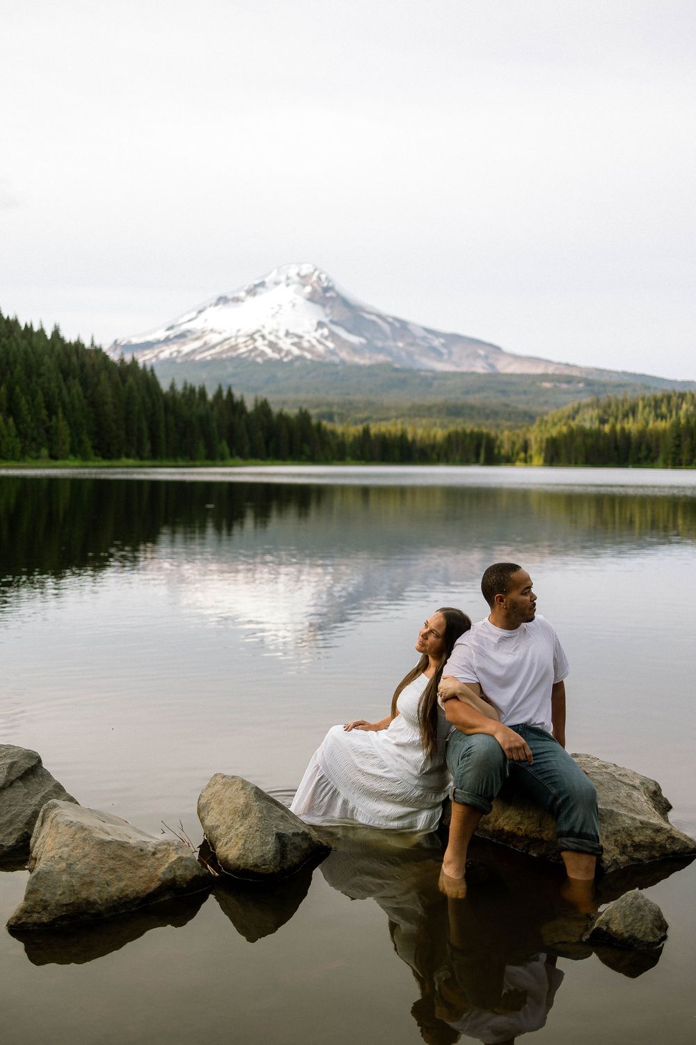 Couple sitting on rocks at Trillium Lake with snow-capped Mt. Hood reflection, romantic outdoor portrait in Oregon.