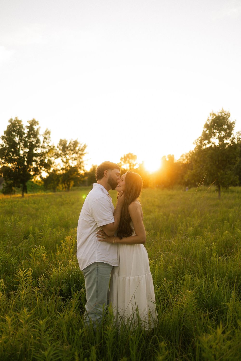 Couple sharing a romantic kiss in a lush green field during golden sunset, woman in white dress, man in white shirt.