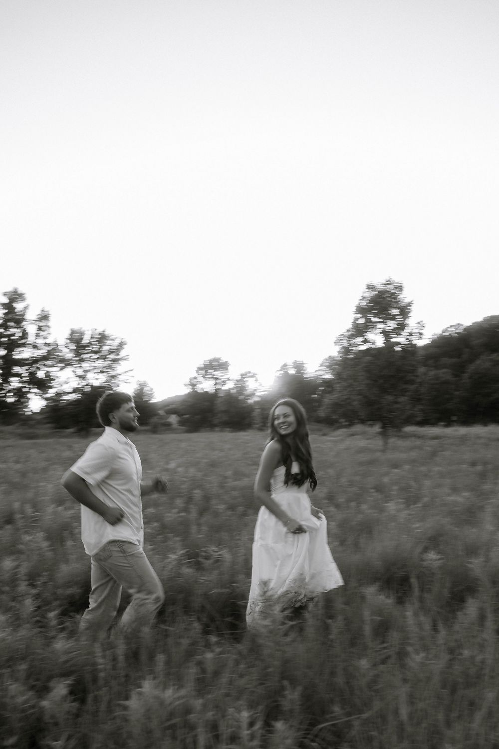 Black and white photo of a couple walking through a meadow, woman in white dress smiling, trees in background.