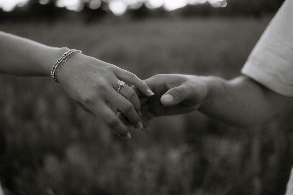 Black and white photo of two people gently holding hands, one wearing a ring and bracelet, in a field.