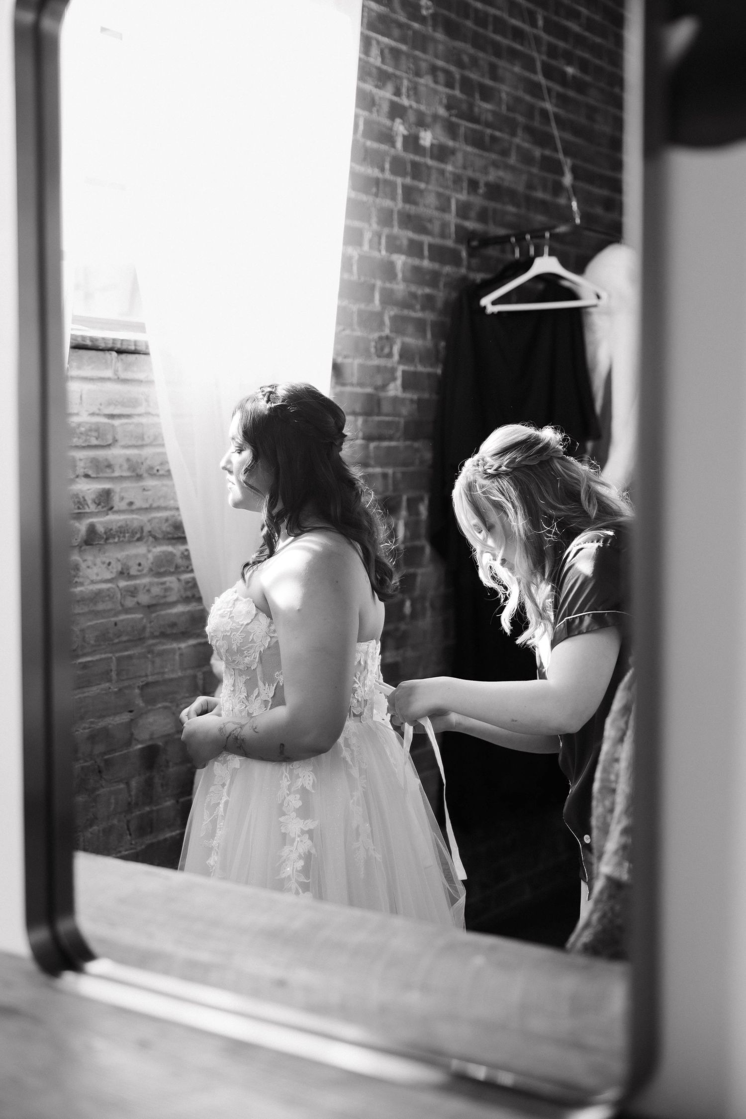 Black and white photo of wedding preparations showing bridal gowns and dresses hanging against exposed brick walls.