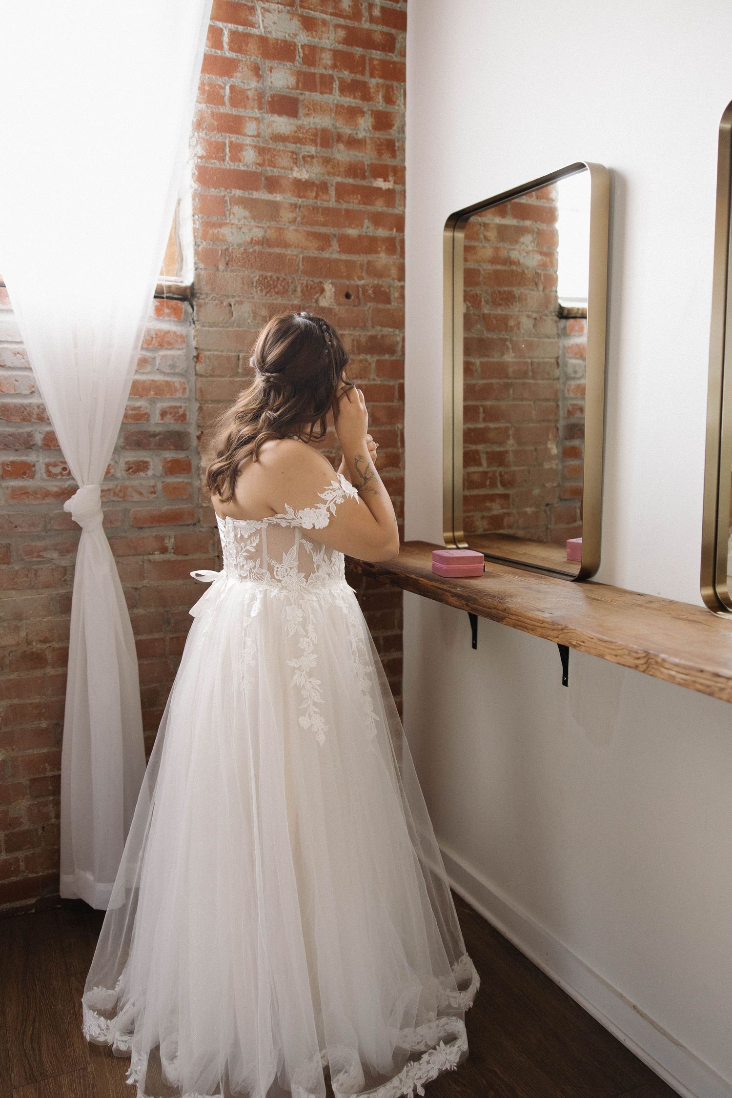 A bride in a white tulle wedding gown stands by a brick wall with mirrors in a getting ready room.