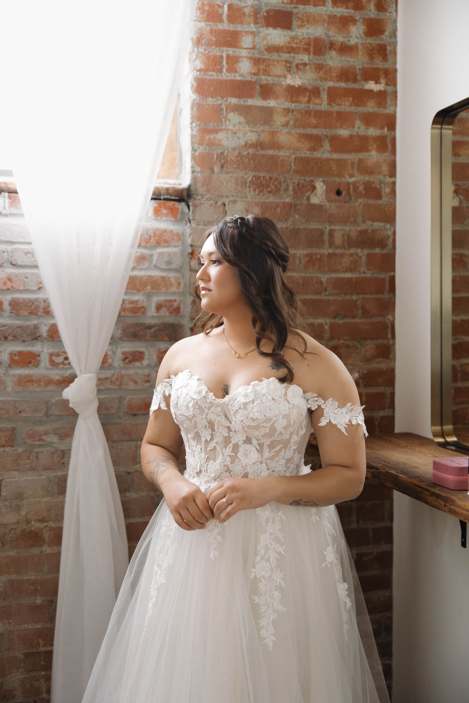 A beautiful wedding dress with delicate lace details and off-shoulder design hangs in a rustic brick-walled room.