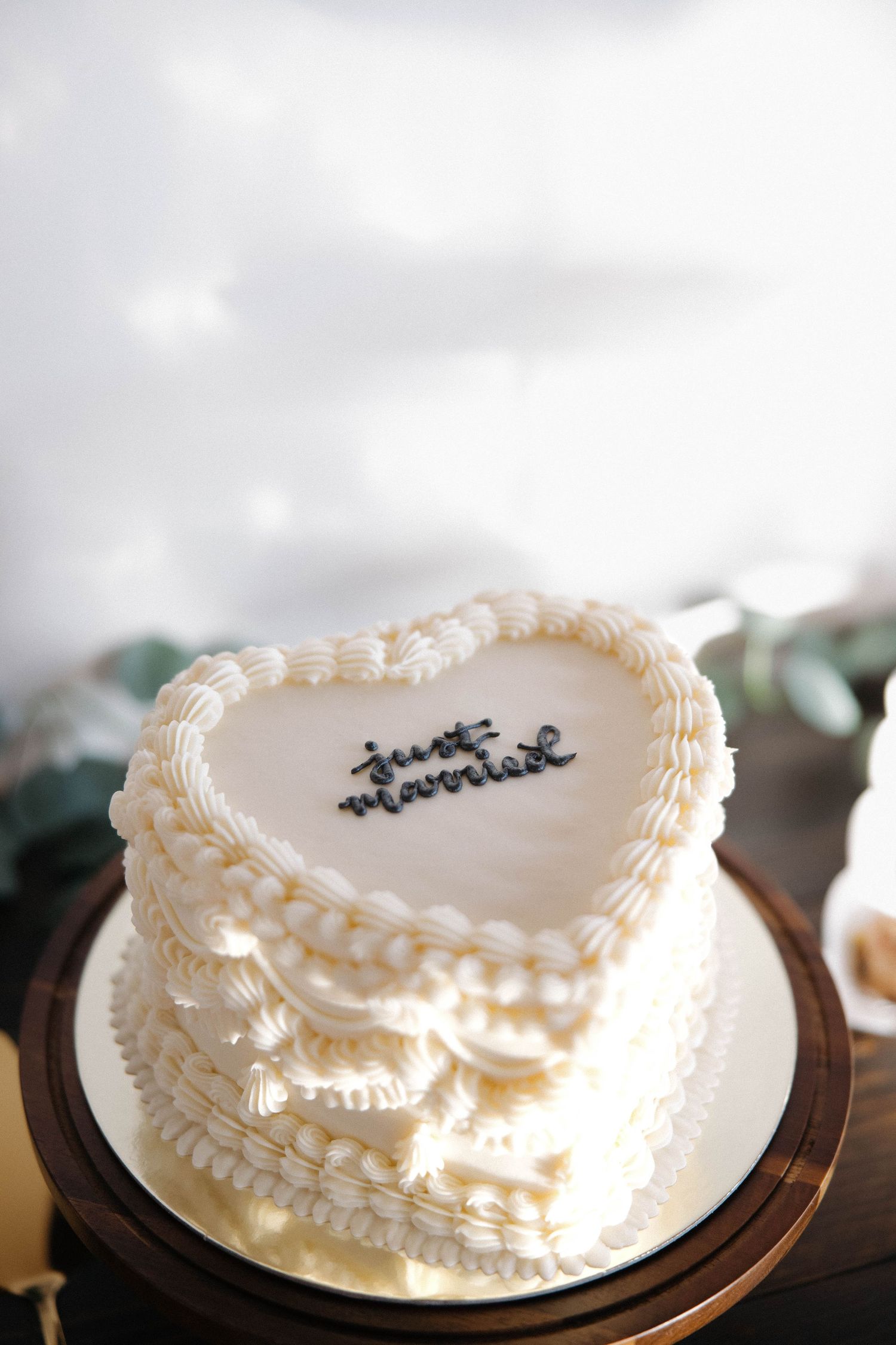 Heart-shaped white buttercream cake with ruffled decorative piping and black text writing displayed on wooden stand.