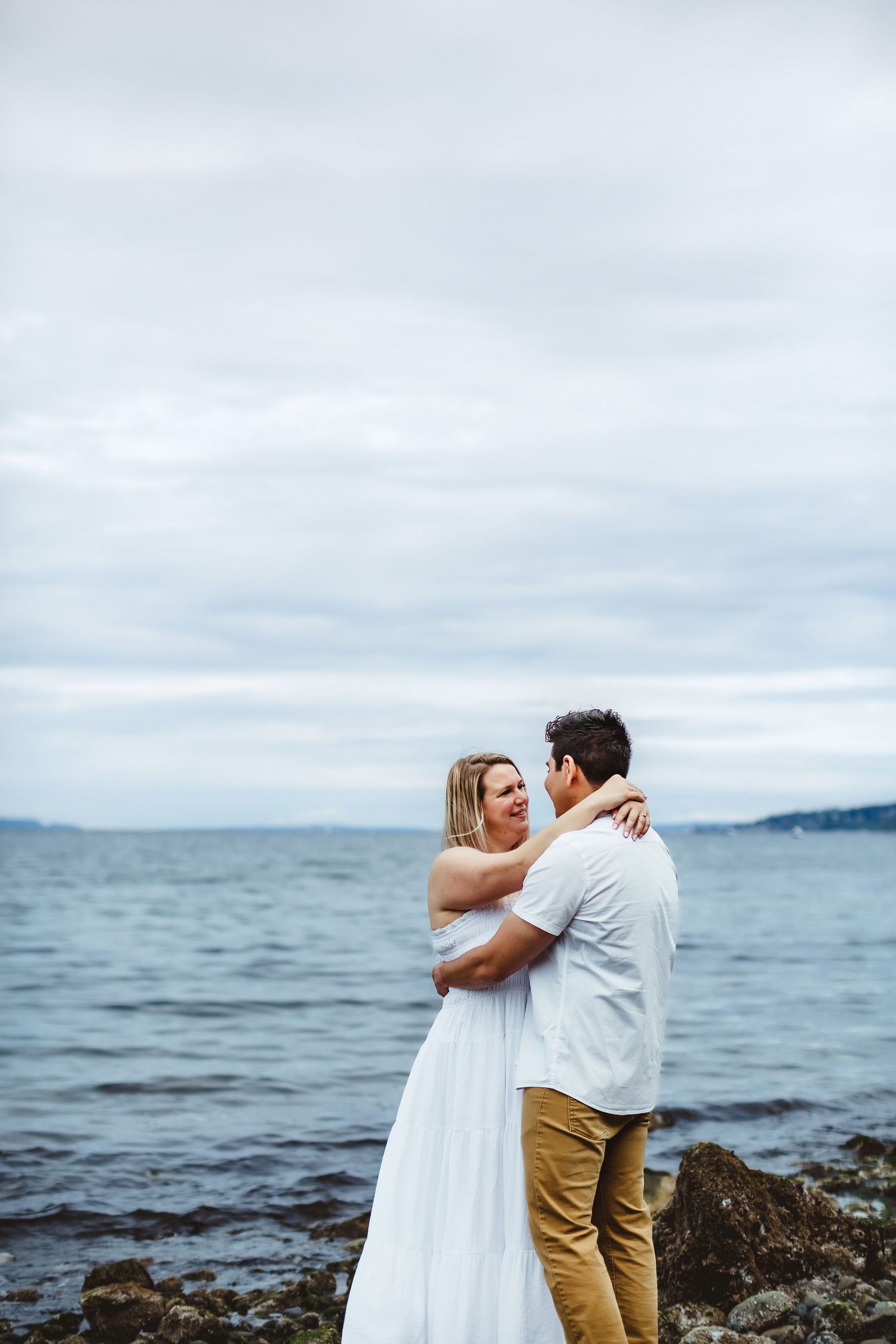 A romantic coastal photoshoot features a couple embracing by the ocean under cloudy skies.