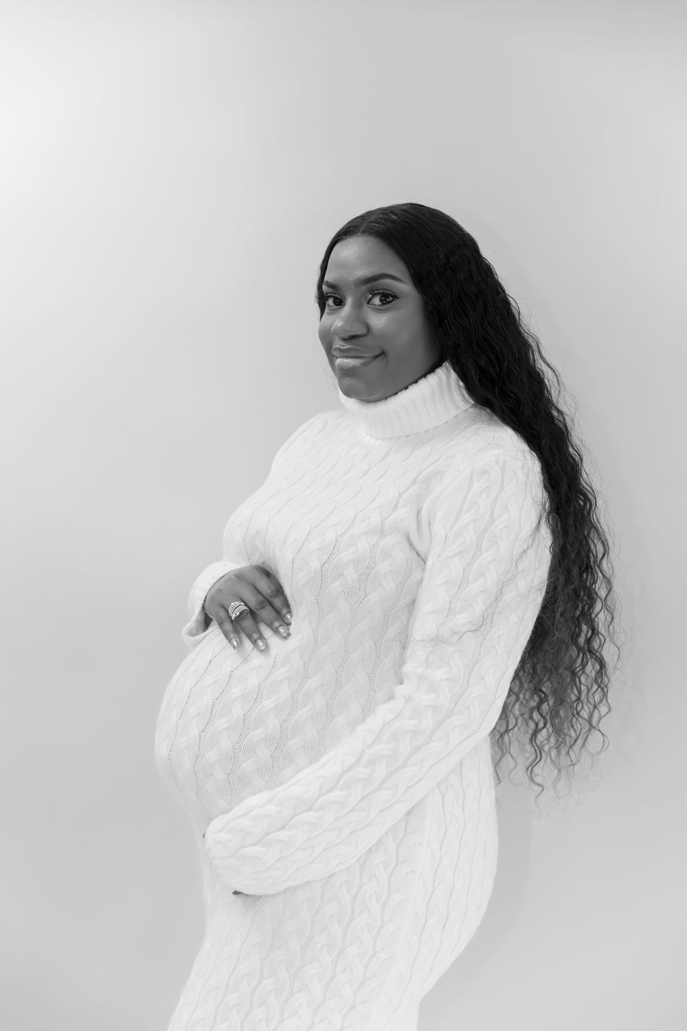 Black and white portrait of a pregnant woman in a white cable-knit sweater dress holding her belly.