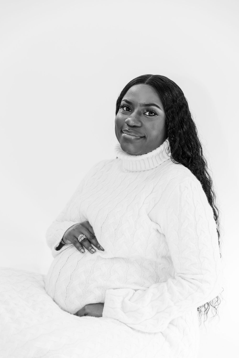 Black and white portrait of a smiling pregnant woman in white cable-knit dress sitting gracefully.