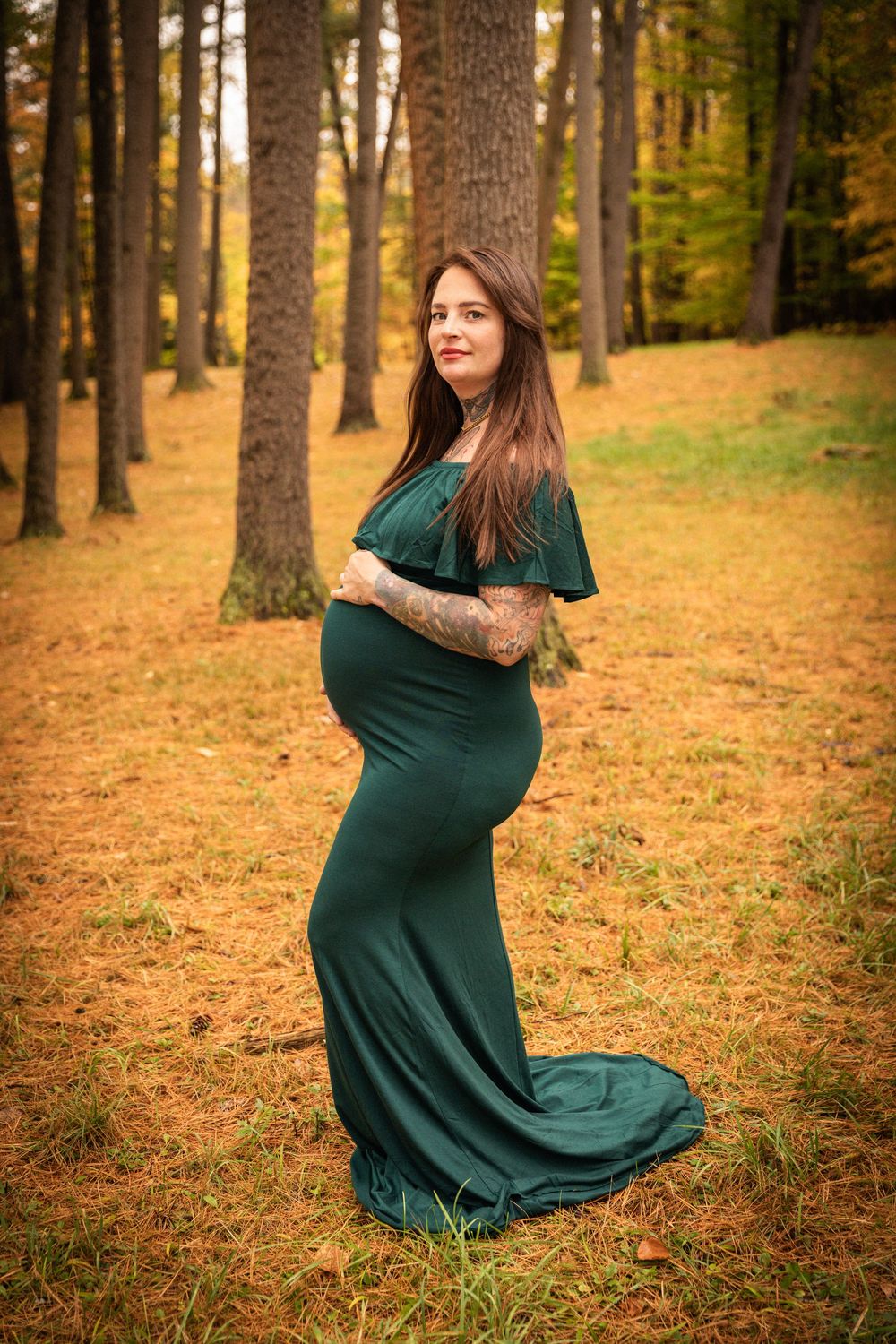 Pregnant woman in elegant emerald green off-shoulder gown poses in an autumn forest setting.