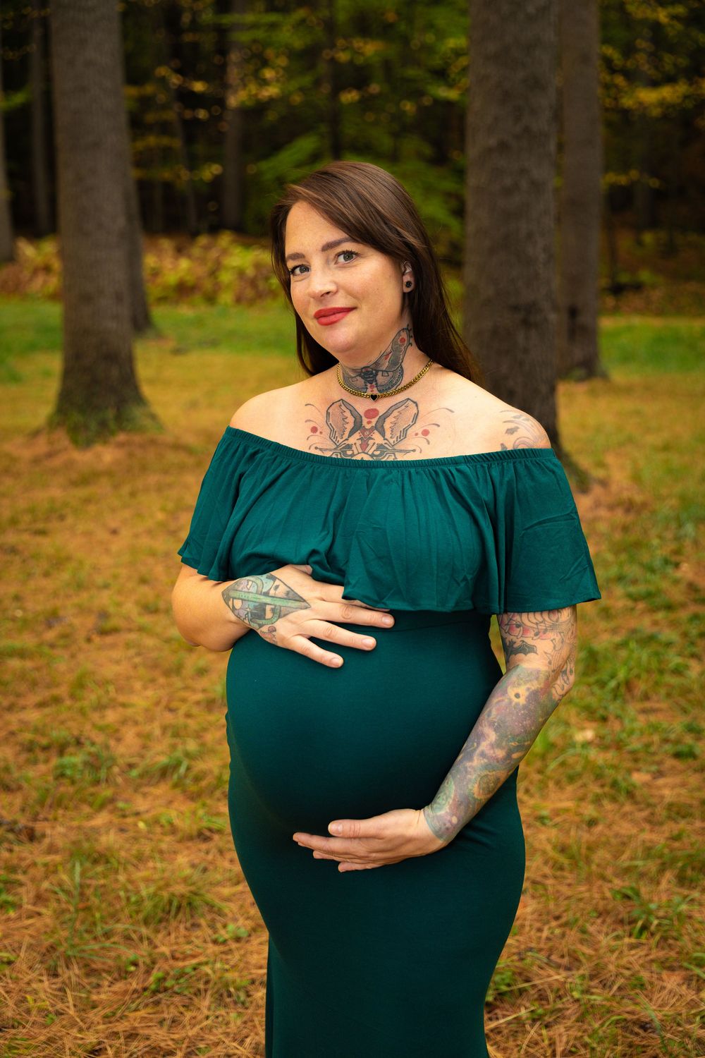 Tattooed pregnant woman in teal off-shoulder dress holds her belly in an autumn forest.