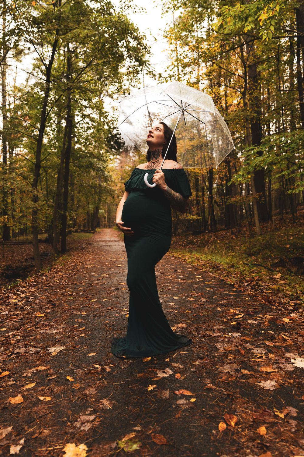 Pregnant woman in green gown holding clear umbrella on autumn forest path with fallen leaves.