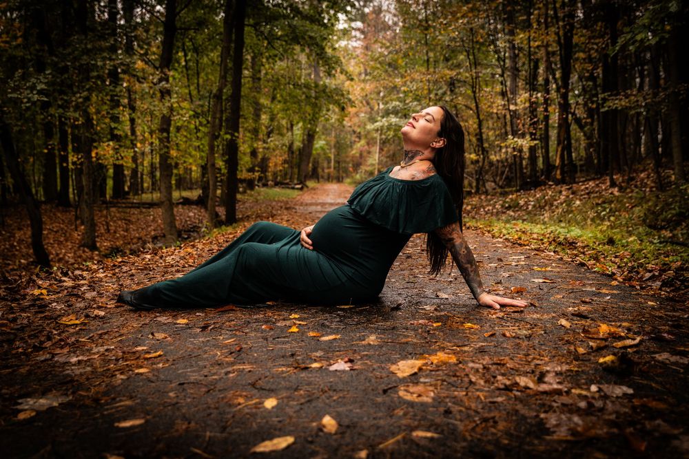 Pregnant woman in green dress sitting on autumn forest path, surrounded by fallen leaves and trees.