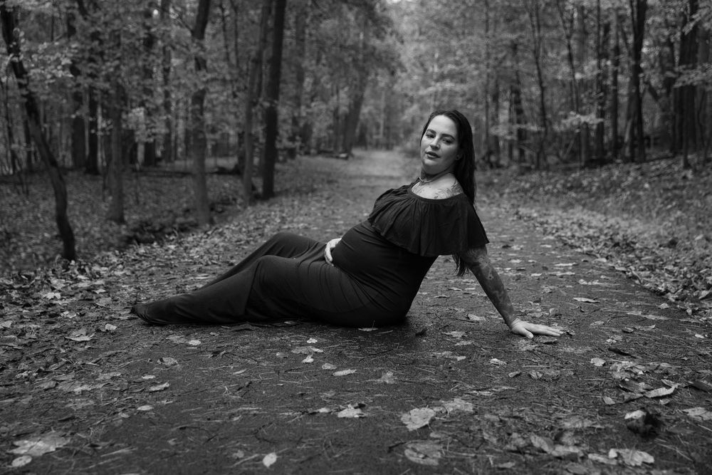 Black and white photo of a woman in a dark dress posing gracefully on a forest path surrounded by autumn leaves for a dreamy maternity shoot