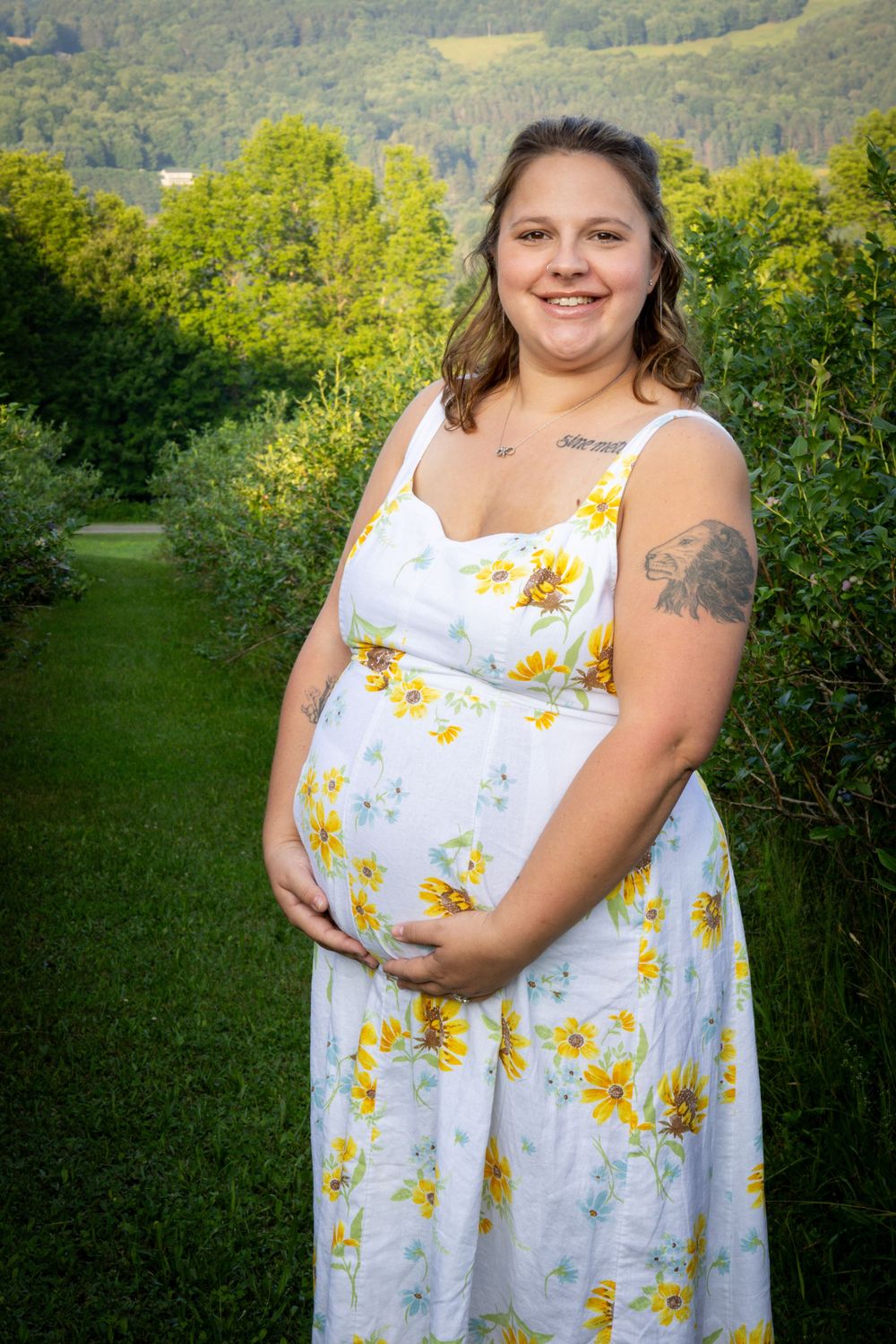 Smiling pregnant woman in white sunflower dress posing outdoors in lush green garden with rolling hills in background.