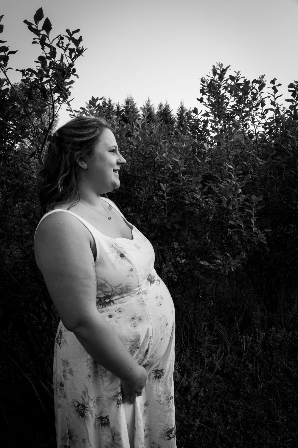 Black and white photo of a pregnant woman in a floral dress standing outdoors among lush green bushes.