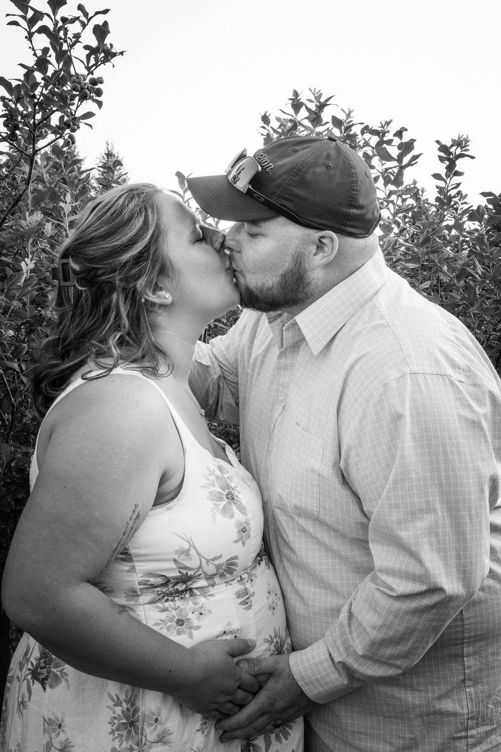 Black and white photo of a couple sharing a kiss outdoors, with the woman in a floral dress cradling her baby bump.