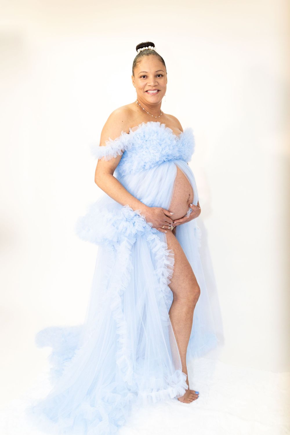 Pregnant woman in elegant light blue tulle off-shoulder maternity gown posing gracefully against white background.