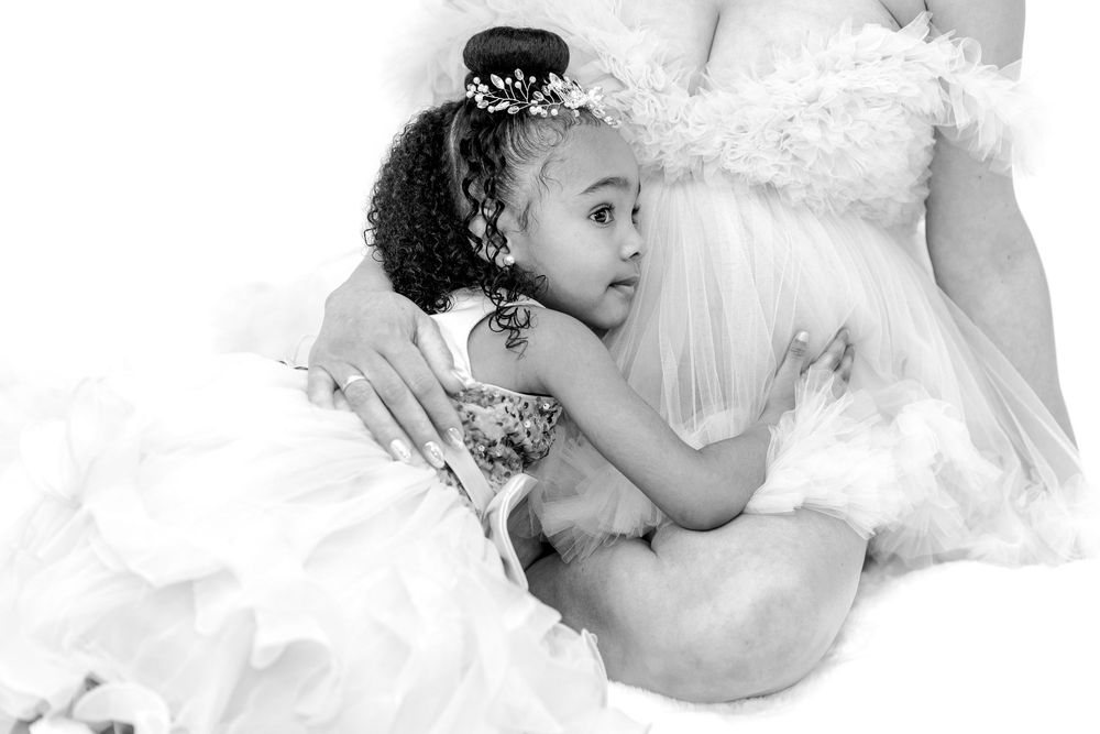 Black and white photo of a young girl in a white dress hugging a mom in a flowing maternity gown.