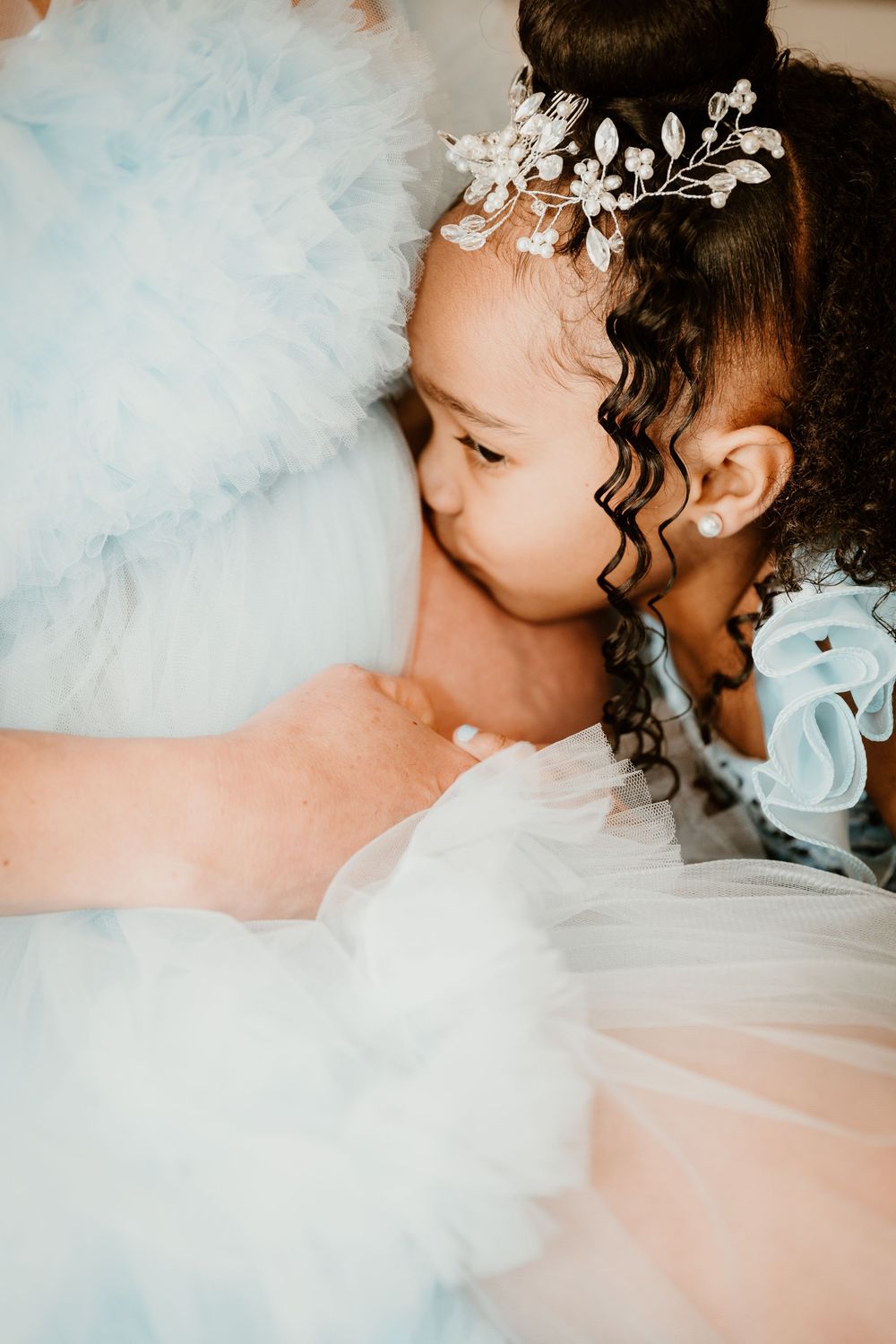 Young girl in light blue tulle dress with floral hair accessory hugging her mother's growing pregnant belly.