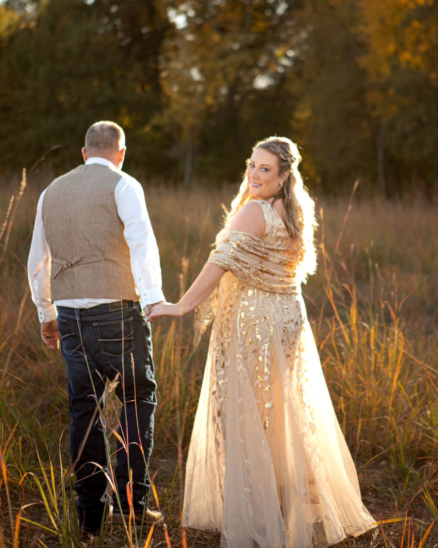 Wedding couple walking through golden grass at sunset wearing formal attire.