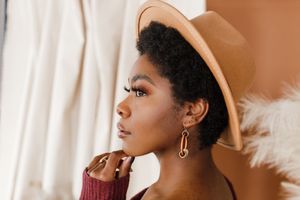 A person in a beige fedora hat and gold hoop earrings looks upward with a contemplative expression.