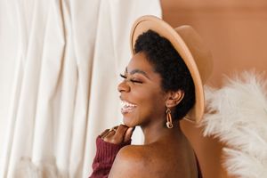 Three diverse women laughing and smiling together while sitting on a couch in a bright sunlit room.