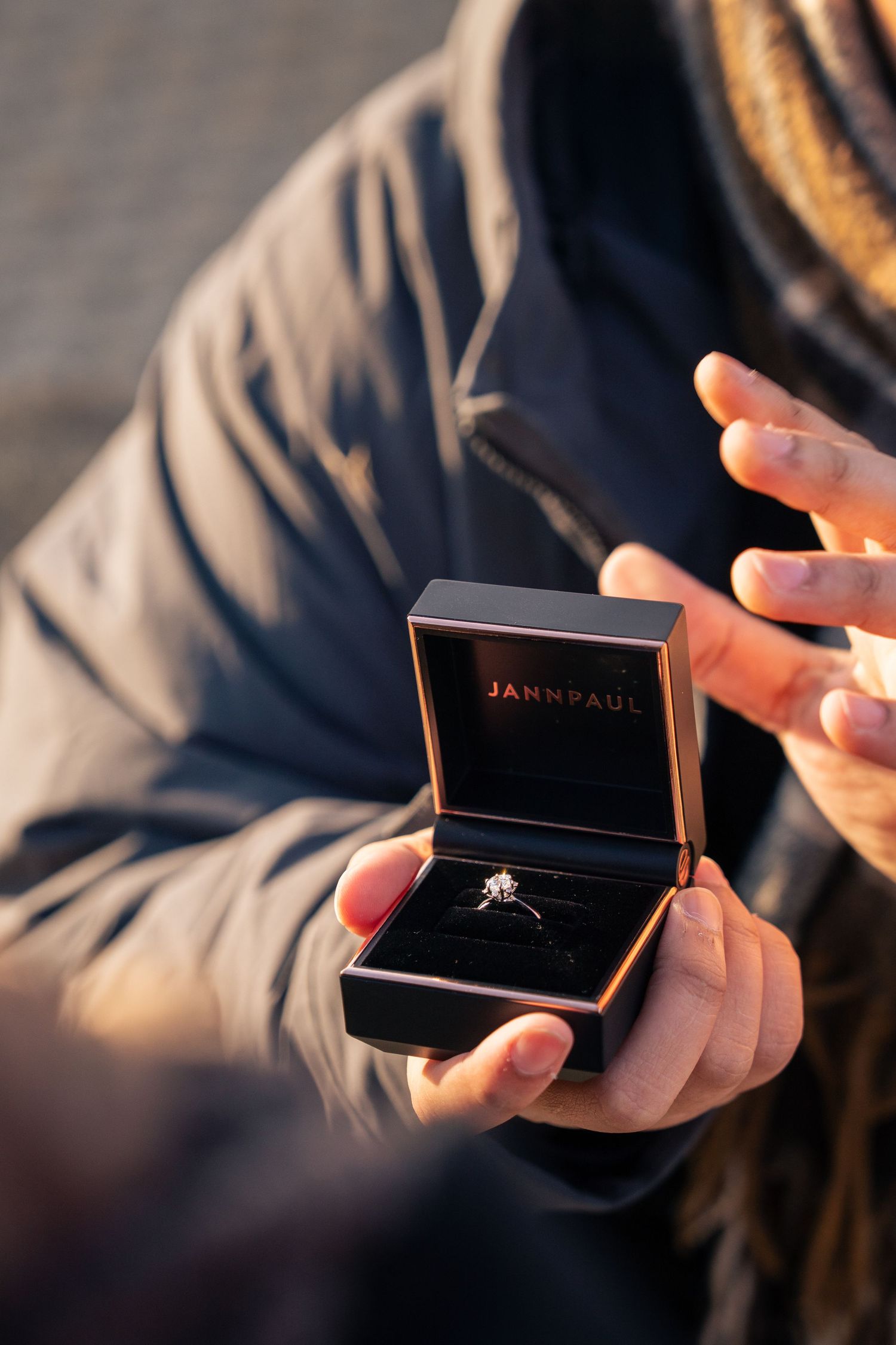 Hands holding open a black Jann Paul jewelry box containing a sparkling diamond engagement ring.