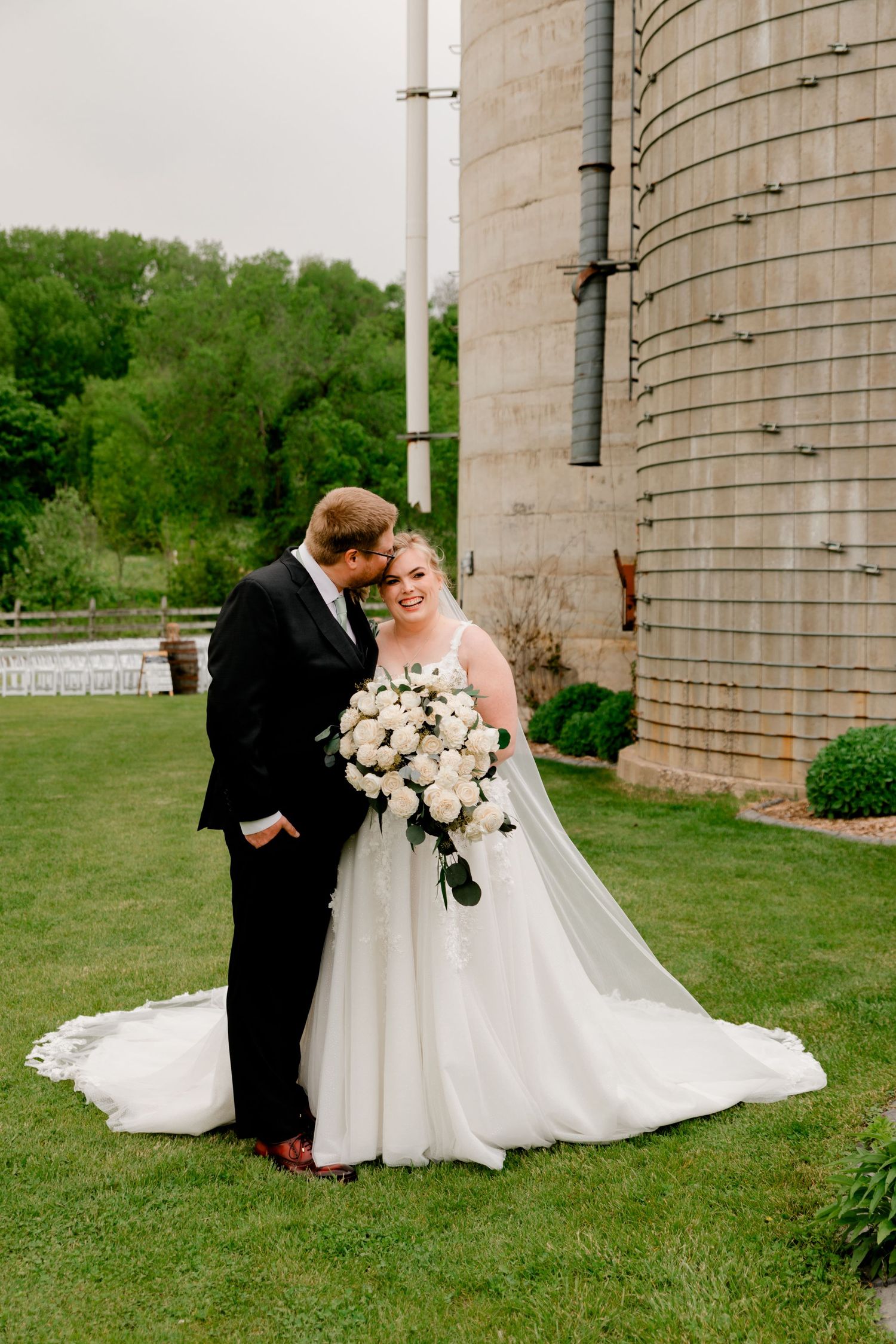 Bride and groom pose together near a rustic grain silo, bride holding a white rose bouquet with flowing veil.