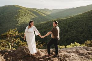 Couple holding hands on rocky mountain overlook, woman in white dress, lush green mountains in background at sunset.