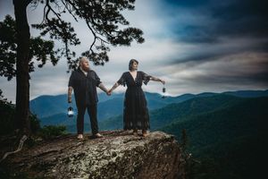 Couple holding lanterns on mountain cliff edge with scenic blue ridge mountain views and dramatic cloudy sky.