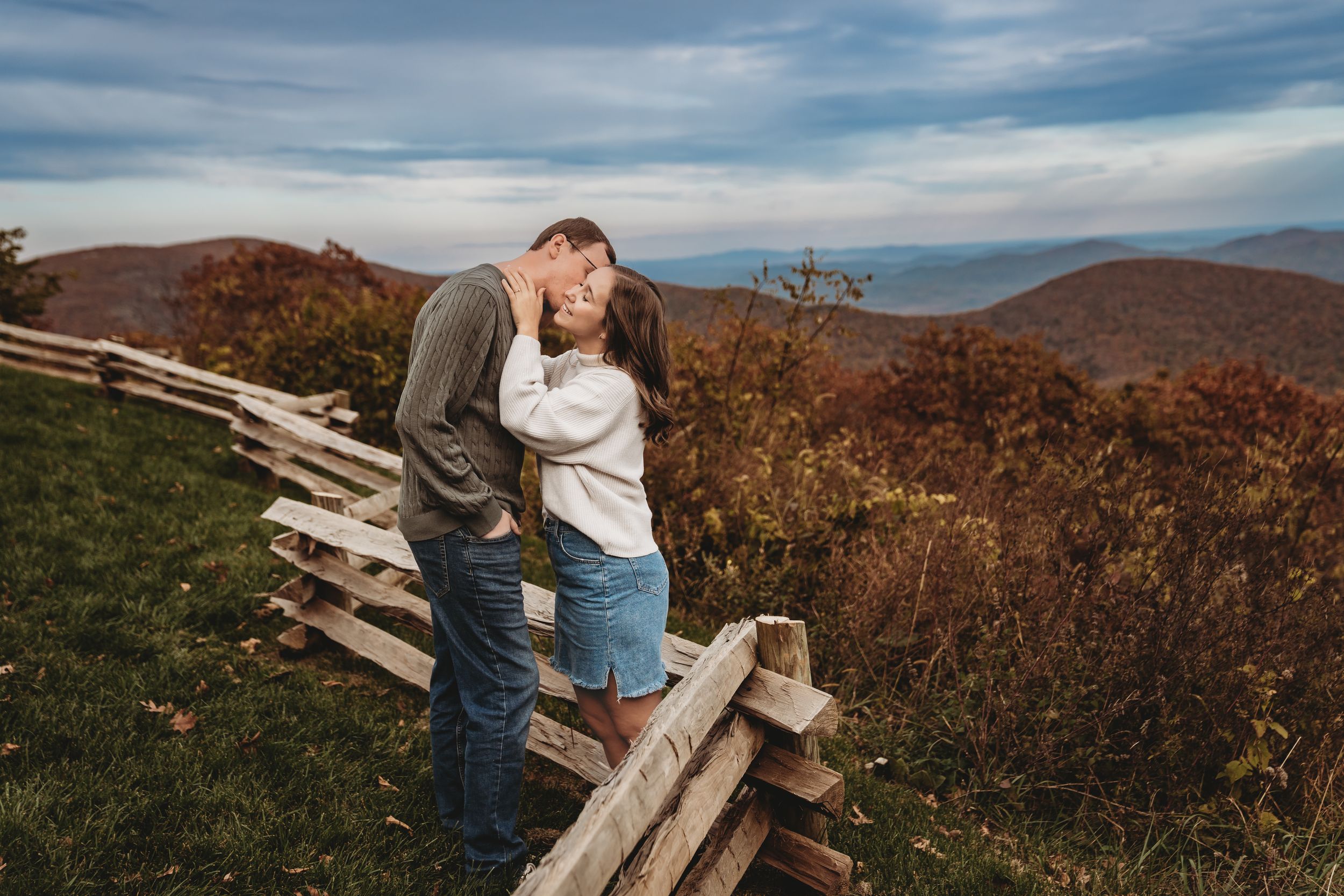 Couple kissing on a mountain overlook in autumn, surrounded by colorful foliage and a rustic wooden fence.