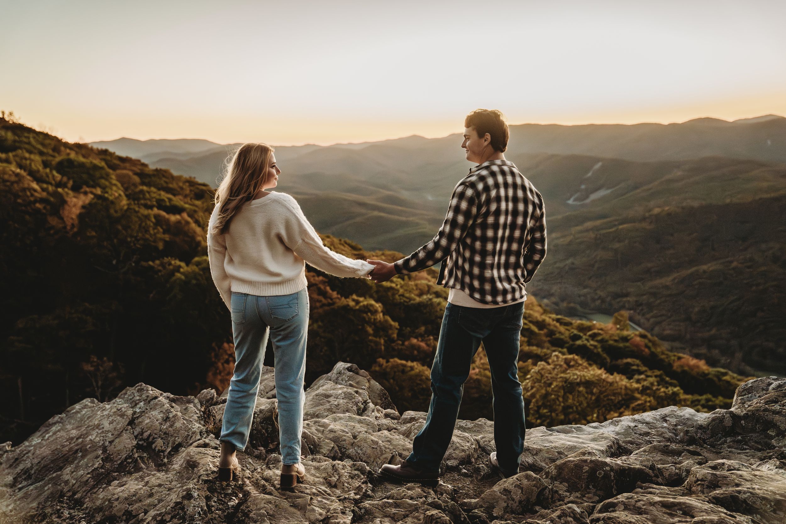 Couple holding hands on rocky mountain overlook at sunset with rolling forested hills in the background.