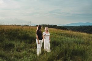 Two women standing in a lush green meadow with mountain views and dramatic sky in the background.