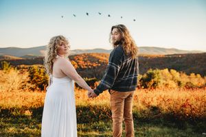 Couple holding hands in autumn field at golden hour with mountain backdrop and birds flying overhead.