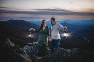 Couple holding lanterns on mountain summit at dusk with blue ridge mountain panorama behind them.