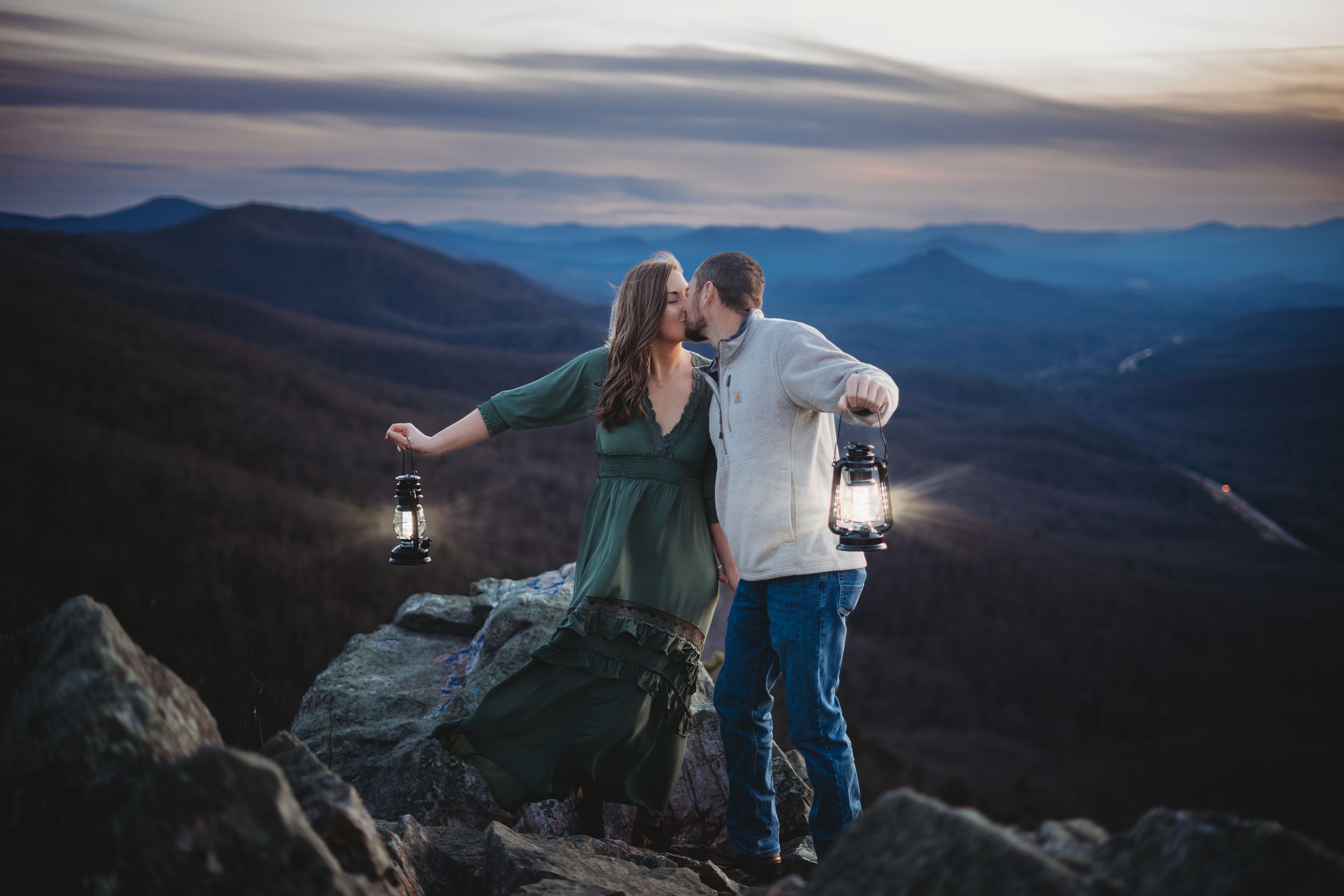 Couple holding lanterns kissing on mountain summit at dusk with blue ridge mountains in background.