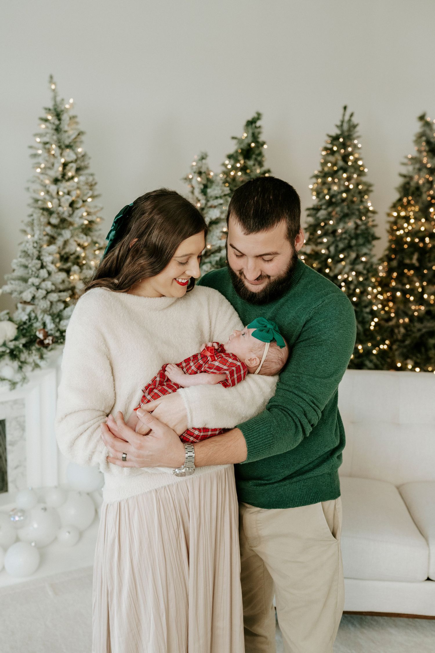 Couple embraces on white couch with decorated Christmas trees glowing in background during holiday photoshoot.