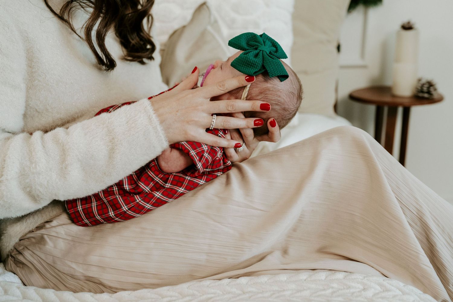 Hands wearing a cozy cream sweater and plaid pajamas hold a festive green bow against white bedding.