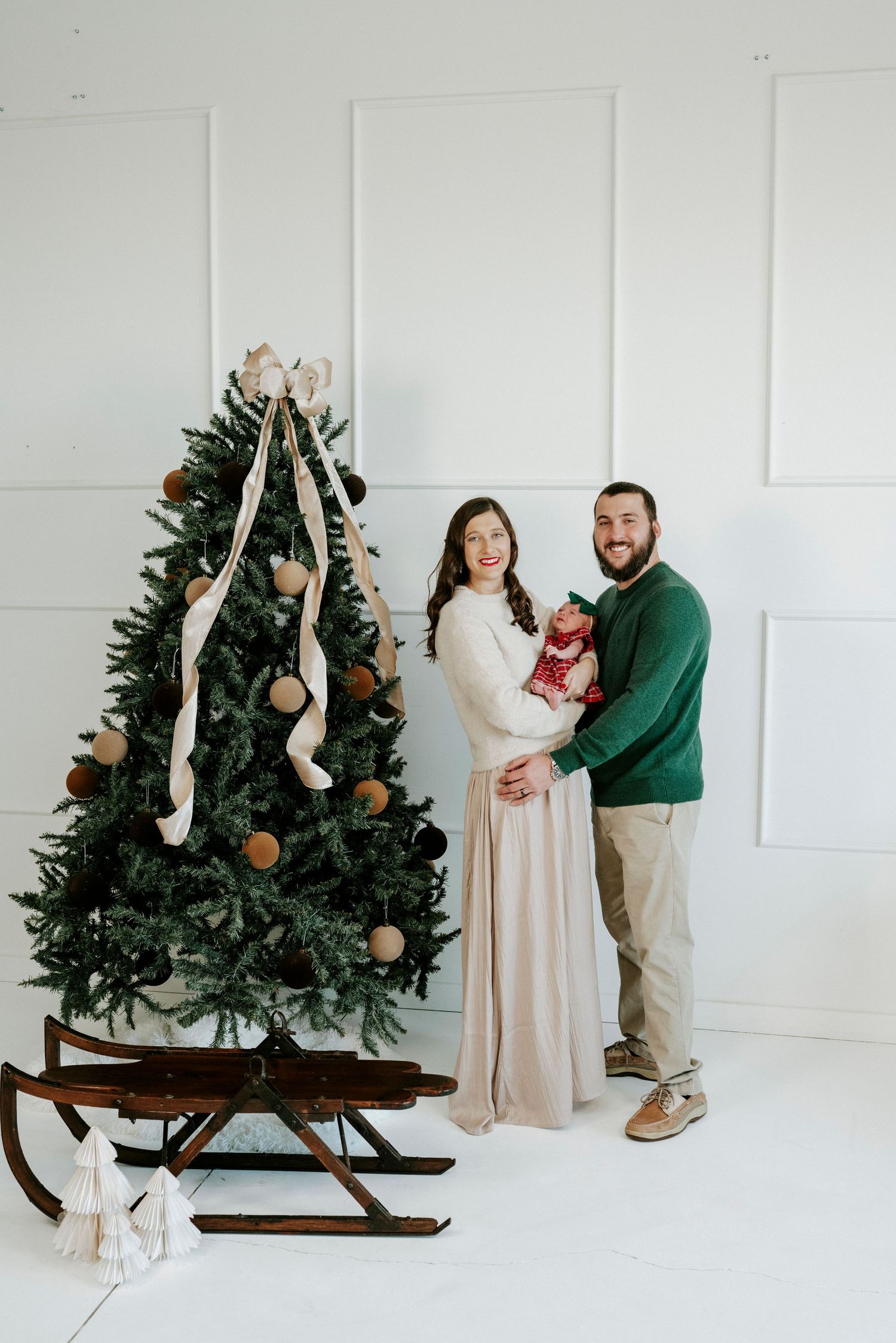 Couple poses playfully with decorated Christmas trees on wooden sleds in bright white studio setting.