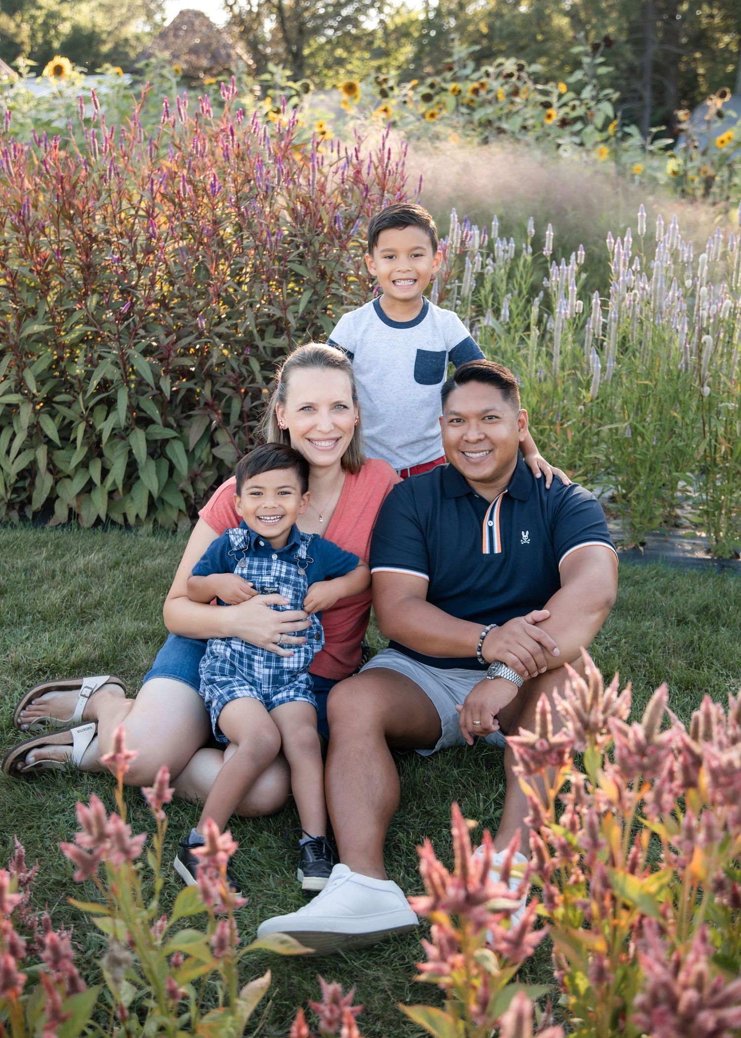 Family members sit together on outdoor furniture surrounded by blooming pink flowers and lush garden vegetation.