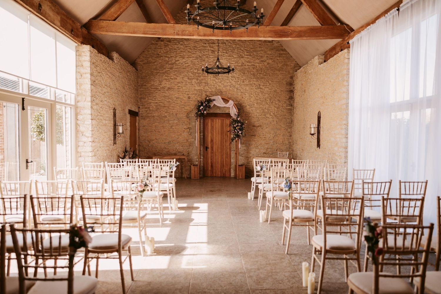 Rustic wedding venue interior with wooden beams, stone walls and chiavari chairs arranged in rows by sunlit windows.