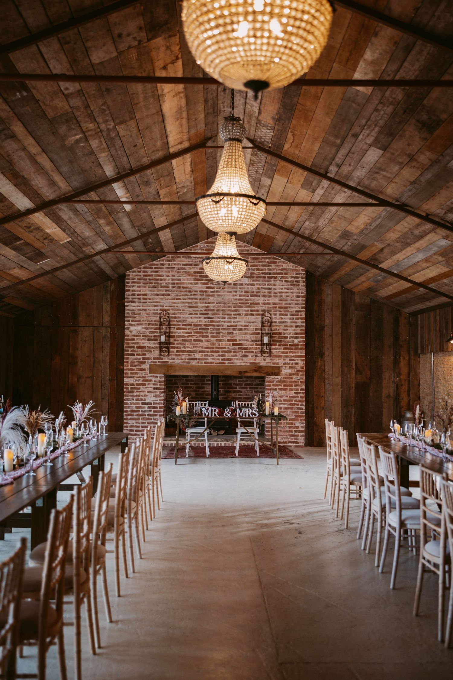 Stratton Court Rustic barn wedding venue interior with wooden beams, crystal chandeliers, and rows of white chairs on concrete floor.