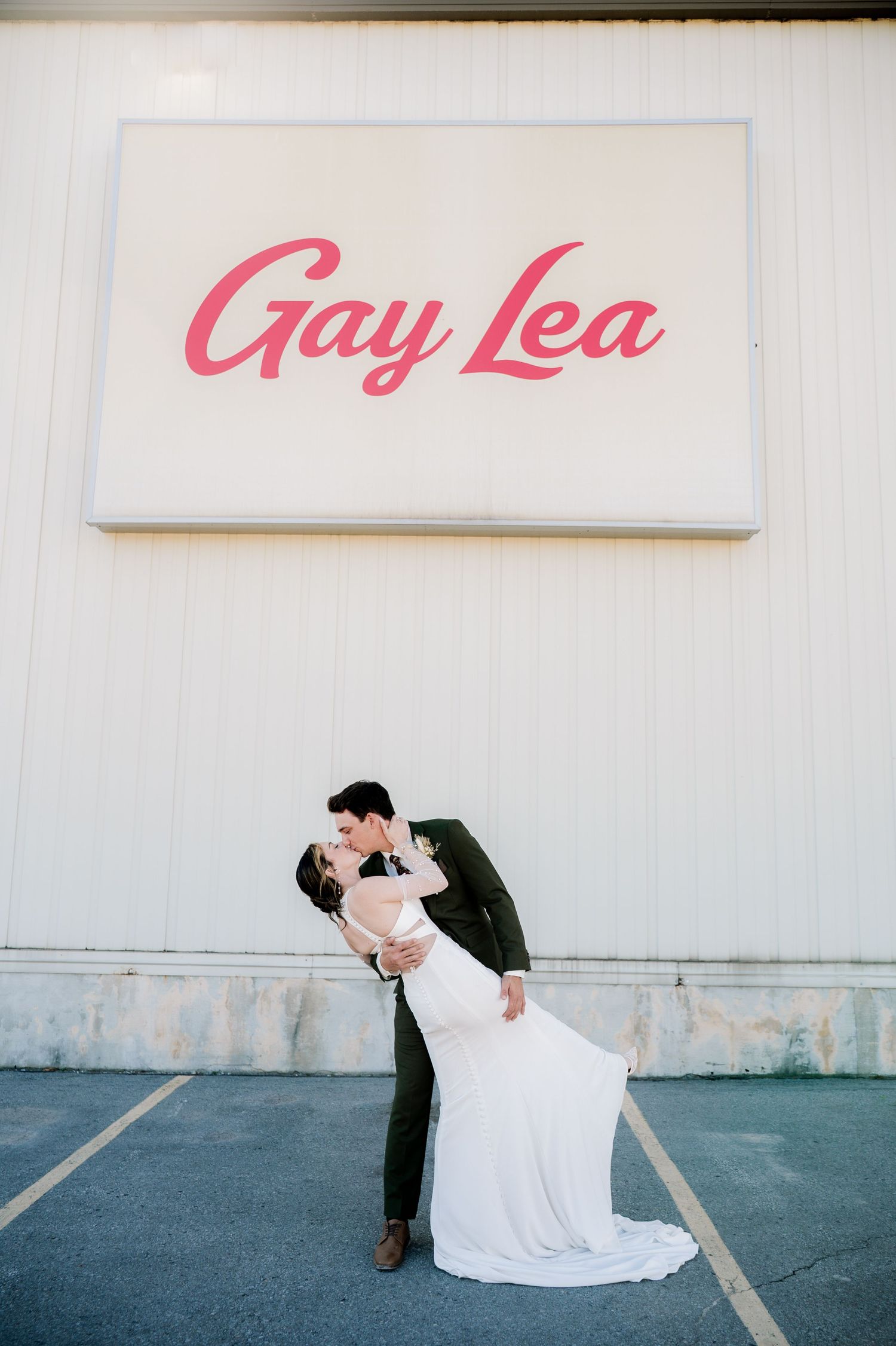 A couple shares a romantic dip kiss under a pink Gay Lea sign on a white wall in a parking lot.