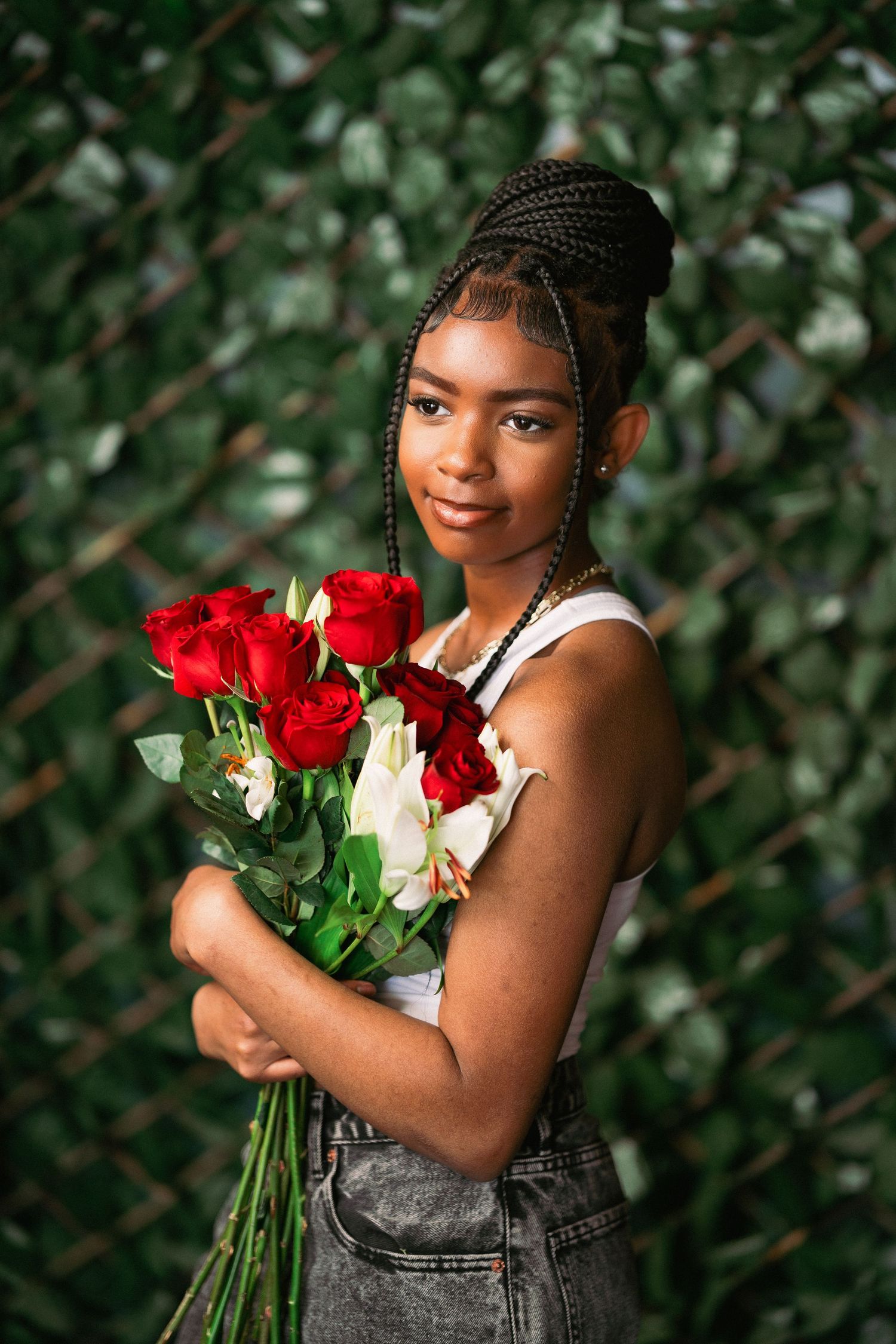 Young woman in denim overalls holds a bouquet of red roses against a green leafy background.