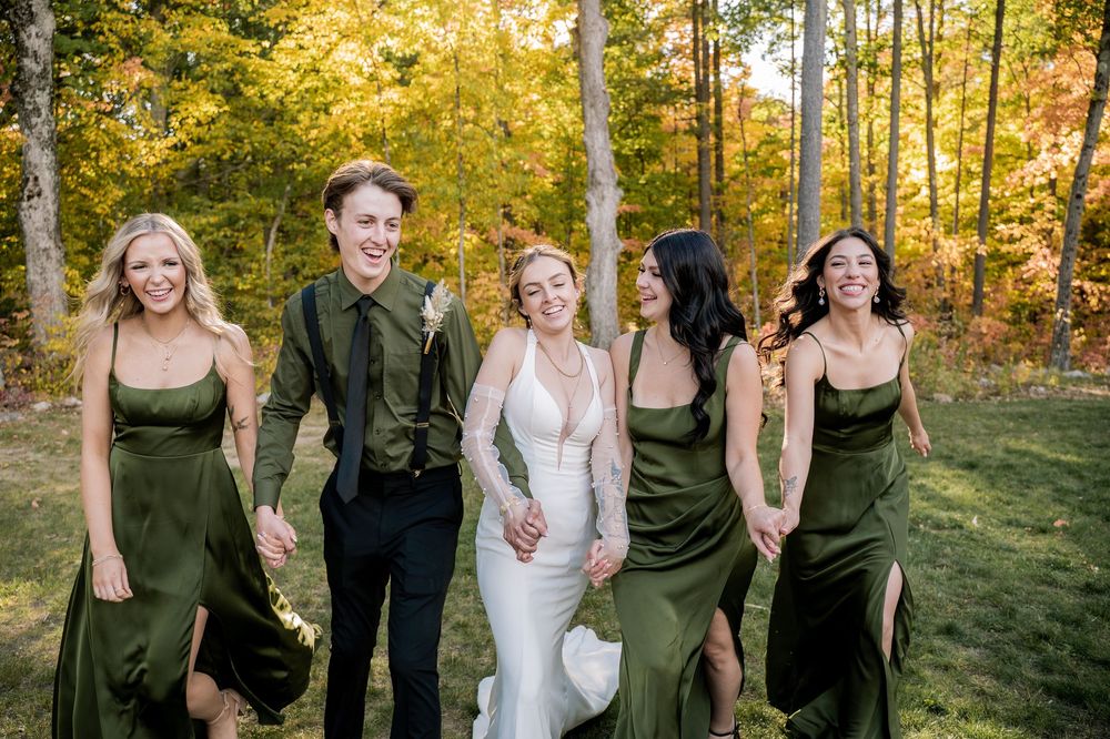 Wedding party poses playfully in a sunlit autumn forest setting with golden foliage in background.