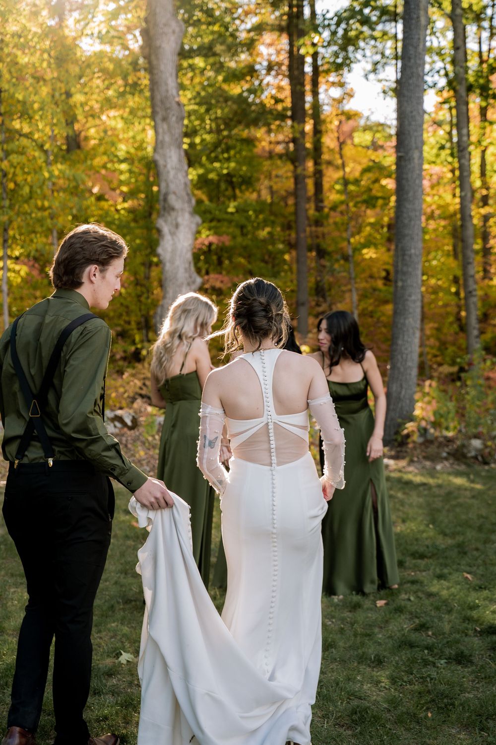 Wedding party stands in a sun-dappled autumn forest with bridesmaids in green dresses and a bride in a white gown.