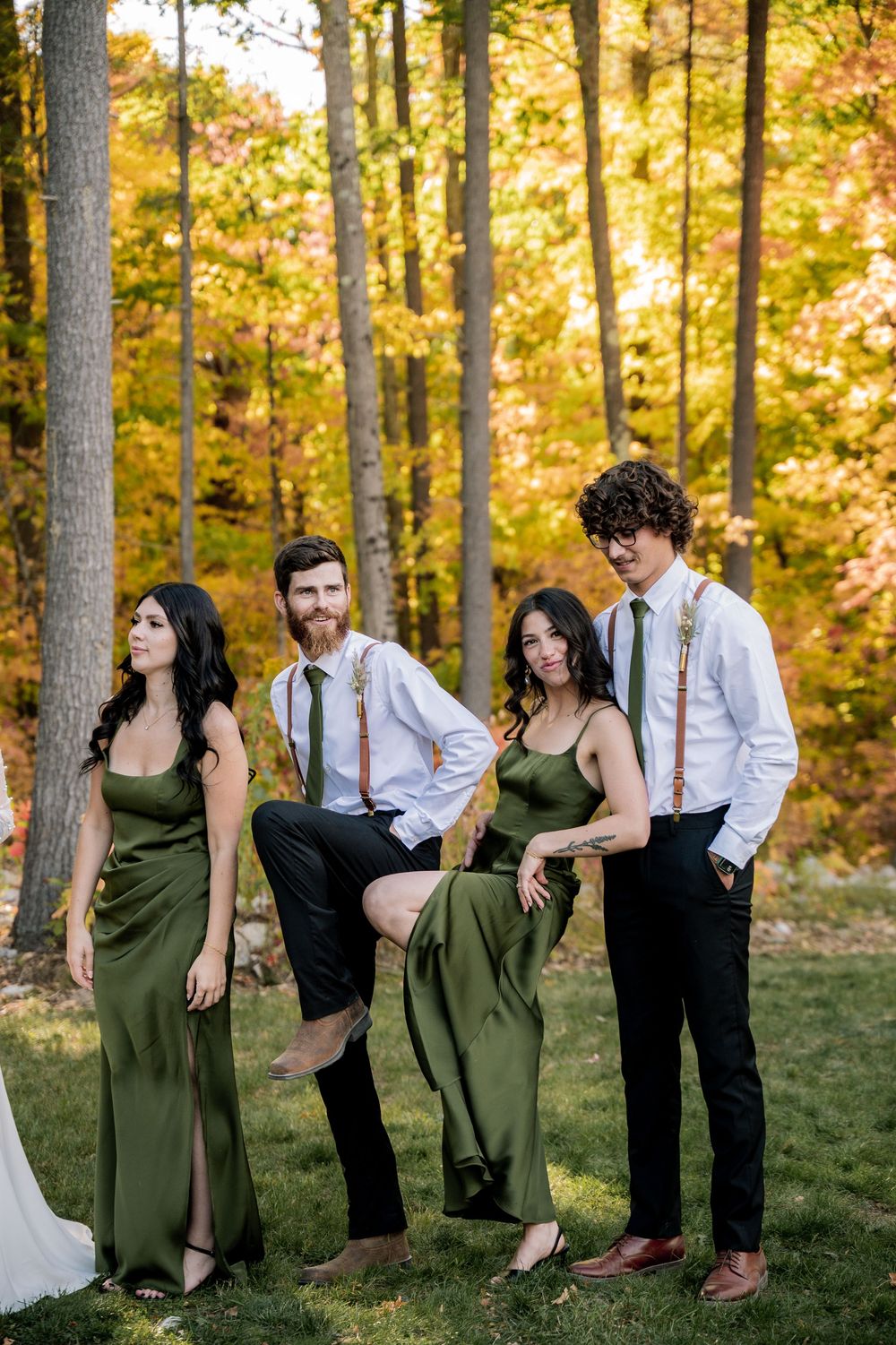 Wedding party poses playfully in a line against autumn forest backdrop with golden foliage and sunlight.